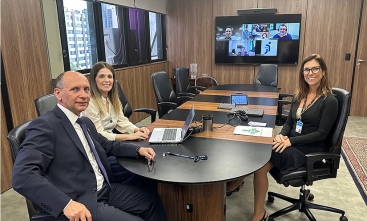 A imagem mostra três pessoas sentadas ao redor de uma mesa de conferência oval em uma sala bem iluminada. Na tela grande ao fundo, aparecem quatro pessoas participando de uma videoconferência. Na mesa, há um laptop aberto, papéis, um telefone e um par de óculos. O ambiente sugere um encontro profissional, possivelmente uma reunião de negócios ou organizacional, com participantes presenciais e virtuais.