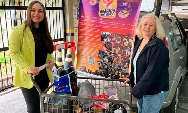 Foto posada de duas mulheres segurando um carrinho carregado de sacolas. Ao fundo está um carro parcialmente coberto por um banner com o texto "Amigos da Sopa" em destaque