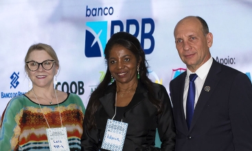  A imagem mostra três pessoas posando para a foto em um evento formal. À esquerda, uma mulher de óculos usa um vestido colorido, ao centro, uma mulher de cabelo preto veste um traje formal preto, e à direita, um homem de terno e gravata azul