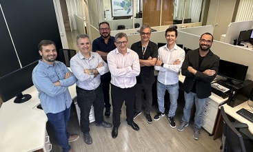 A foto mostra um grupo de sete homens em pé, sorrindo e de braços cruzados, posando para a câmera em um ambiente de escritório. Todos estão em um local bem iluminado, com divisórias e várias mesas com computadores ao fundo.