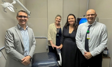 Foto posada de quatro pessoas em uma sala com alguns equipamentos eletrônicos ao fundo e uma cadeira reclinável de consultório médico