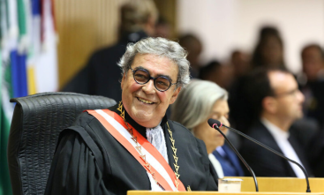 A foto mostra uma pessoa com cerca de 60 anos, cabelos grisalhos, sorrindo, e vestindo uma toga judicial. Ela está sentada em um ambiente de tribunal.