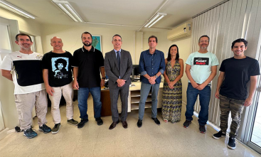 Na foto, sete pessoas estão lado a lado, posando para a câmera em um ambiente fechado, como um escritório. Três homens estão à esquerda, dois deles com camisetas informais e o terceiro vestindo uma camisa preta. Ao centro, um homem de terno cinza, seguido por três pessoas à direita: um homem de camisa azul, uma mulher de vestido floral e dois homens em trajes casuais. Ao fundo, há uma mesa com alguns objetos de  escritório e janelas com persianas.