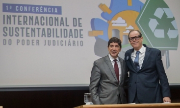 A imagem mostra dois homens de terno posando para uma foto, sorrindo e de frente para a câmera. Eles estão em pé, lado a lado, em frente a um painel que apresenta o título "1ª Conferência Internacional de Sustentabilidade do Poder Judiciário". No fundo, há ilustrações coloridas, incluindo um símbolo de reciclagem e imagens que remetem à natureza e sustentabilidade.