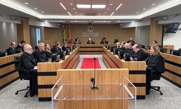 Fotografia da sala de sessões do Tribunal Pleno, com mesa em "U", juízes em togas e bandeiras ao fundo