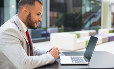 Fotografia ilustrativa de homem negro sorridente vestido formalmente olhando para tela de um notebook ao ar livre