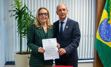 A imagem retrata duas pessoas em pé, lado a lado, segurando um documento. À esquerda, uma mulher vestida com um blazer xadrez verde e preto, óculos de armação grossa e cabelo loiro solto, sorri para a câmera. À direita, um homem de terno escuro, gravata e com expressão mais séria, também olha diretamente para a câmera. Ao fundo, há uma bandeira do Brasil, uma planta em vaso e uma parede com persianas