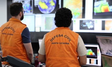 Fotografia de dois agentes da Defesa Civil monitorando telas de clima. Ambos estão de costas para a câmera.