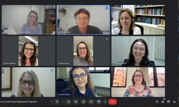 Esta é uma imagem de uma reunião virtual realizada por videoconferência, onde nove pessoas aparecem em suas respectivas janelas de vídeo, organizadas em uma grade. Todas as pessoas estão sorrindo, e há uma variedade de fundos, como estantes de livros e janelas, que mostram diferentes ambientes de trabalho. Os participantes parecem estar concentrados na reunião e estão todos vestidos de maneira profissional.