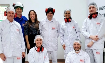 A foto mostra um grupo de cinco homens e três mulheres, sorridentes, a maioria vestindo uniformes brancos com o logotipo "Tirol" e usando equipamentos de proteção, como redes para cabelo e protetores auriculares vermelhos, típicos de um ambiente de produção ou fábrica. 