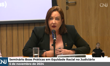 Foto de uma mulher branca de cabelo castanho até os ombros falando ao microfone. Ela veste um blazer preto e uma peça bege coberta pelas mãos. Em uma barra abaixo está escrito "Seminário Boas Práticas em Equidade Racial no Judiciário", ao lado o logo do CNJ e abaixo "5 de novembro de 2024"