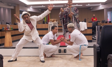 A imagem mostra uma apresentação cultural em um auditório. Em destaque, um homem vestido com um traje branco e chapéu executa uma expressão corporal estilizada, enquanto dois outros, com uniformes brancos de capoeira, estão agachados no centro realizando um cumprimento típico dessa arte. Ao fundo, uma pessoa toca berimbau, e há outros elementos tradicionais, como tambores, além da decoração do auditório com madeira e bandeiras