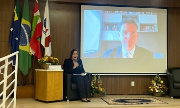 A imagem mostra um palco onde uma mulher está sentada à esquerda, de frente para uma grande tela que exibe outra pessoa. A pessoa sentada está segurando um tablet. Na tela, um homem é mostrado da cintura para cima, em uma videochamada. O fundo da sala inclui várias bandeiras e arranjos florais em ambos os lados da área do palco. 