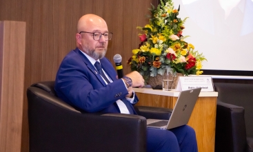 Homem de terno azul usando óculos sentado em poltrona, com microfone na mão e notebook no colo