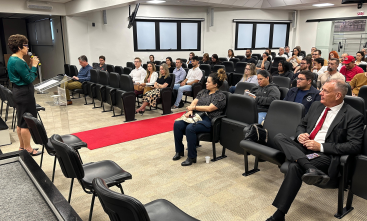 Fotografia de um auditório no qual uma mulher fala em pé em um púlpito à frente de um público que está sentado.