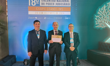 Três homens de terno escuro com um banner iluminado ao fundo onde se lê 18º Encontro Nacional do Poder Judiciário. À esquerda está um homem amarelo (de traços extremo-asiáticos), de cabelo escuro curto e gravata escura, à direita um homem branco de cabelo escuro curto e gravata quadriculada, ao meio um homem branco, calvo, de gravata vermelha, mostrando um certificado