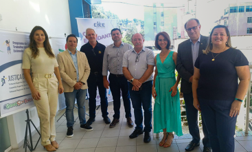 Grupo de oito pessoas em um espaço aberto, ao lado de banners de eventos, com ambiente iluminado e prédios ao fundo. Todos sorriem para a câmera.