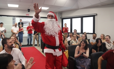 Homem vestido de Papai Noel desce uma rampa dentro de um auditório, sendo aplaudido pelo público presente