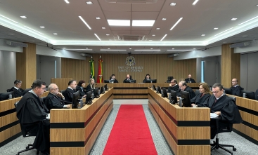 Fotografia em plano aberto da sala de sessão do Tribunal Pleno do TRT-SC. Ao centro, um corredor com um tapete na cor vermelha se estende até o fundo, onde há uma bancada onde está sentado o presidente da corte. Nas laterais direita e esquerda, há duas fileiras de postos de trabalho individuais, onde estão sentados os desembargadores.