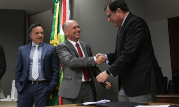 Dois homens de terno apertam as mãos sorrindo, em um ambiente formal com bandeiras ao fundo. Um terceiro homem ao lado, também de terno, observa a interação.