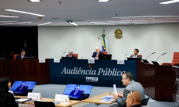 Plenário onde se encontram entre sete pessoas. Três homens se encontram ao centro da mesa na qual está escrito "audiência pública" e possui o logo do STF.