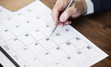 Fotografia em plano detalhe de uma mão segurando uma caneta apontando para uma data em um calendário de papel sobre uma mesa de madeira.