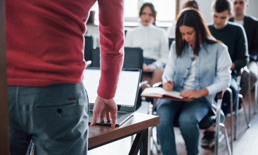 Fotografia de uma sala de aula. Em primeiro plano, um professor de costas está com uma das mãos apoiada sobre uma mesa onde há um notebook aberto. Ao fundo, se vê uma turma de estudantes sentada em cadeiras, fazendo anotações em seus cadernos. 