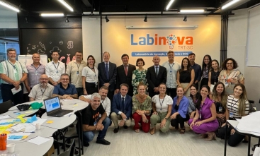 Grupo de 27 pessoas posando juntas em um espaço colaborativo com mesas, post-its e uma placa indicando "Labinova TRT-SC – Laboratório de Inovação".