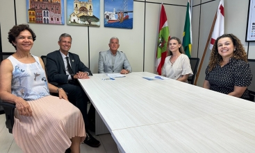 Cinco pessoas estão reunidas em uma sala, sentadas à mesa, com bandeiras e quadros ao fundo. 