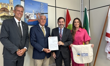 Quatro pessoas posando para foto, duas delas segurando um documento assinado e outra segurando uma bolsa com a logo do Programa "Trabalho Seguro". Ao fundo, três bandeiras