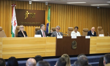 A imagem mostra uma mesa de autoridades no Tribunal Superior do Trabalho (TST), com placas nominais, bandeiras e um público acompanhando a sessão.