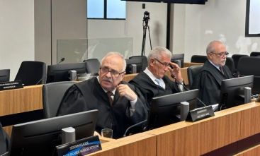 Fotografia da sessão em que houve a decisão. Três homens de toga estão sentados de frente para a tela de seus computadores em ambiente fechado.