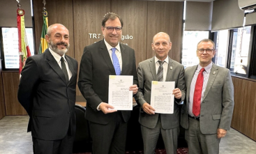 Quatro homens de terno e gravata estão em pé, posando e sorrindo para a foto, em uma sala de reuniões