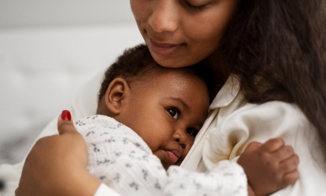 Fotografia ilustrativa de mulher negra com bebê no colo. Ambos vestem roupas claras.