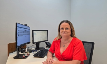 Uma mulher branca de cabelos loiros, usando uma blusa vermelha, está sentada atrás de uma mesa de cor clara, na qual há um computador com dois monitores e um telefone.