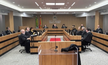 Em um plenário estão sentadas quinze pessoas, sendo três mulheres e doze homens. Eles vestem togas pretas e estão em estações de trabalho posicionadas em quatro fileiras. Ao fundo, estão sentadoes três homens, sendo ao centro, o presidente do TRT-SC.