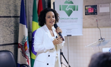 Uma mulher parda de cabelos escuros, curtos e cacheados, trajando um terno branco com calça e camisa brancas, fala em pé ao microfone em um auditório. Ao fundo, um banner onde se lê: Conciliar é Melhor.