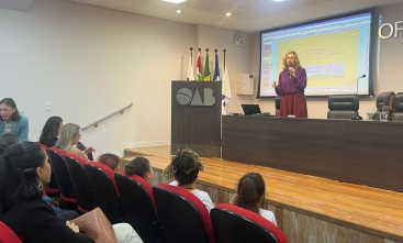 Mulher loira, de blusa rosa em pé diante de um auditório