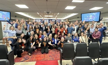Um grupo de aproximadamente 60 pessoas está sorrindo com as mãos para cima, posando para a foto, em uma salão de auditório