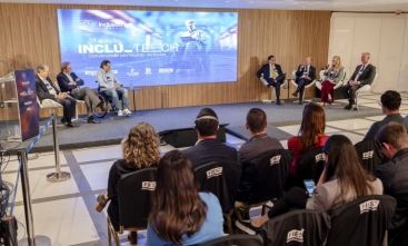 Seis homens e uma mulher estão sentados em um palco de um auditório dando uma palestra. Ao fundo um telão e na frente pessoas estão assistindo