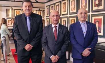 Três homens de terno, em pé, sorriem. Eles estão à frente de uma galeria de quadros
