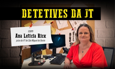 Detetives da JT: Juíza do Trabalho Ana Rick, uma mulher branca de cabelos loiros, usa uma blusa vermelha e está sentada atrás de uma mesa de cor clara, na qual há um computador com dois monitores e um telefone.