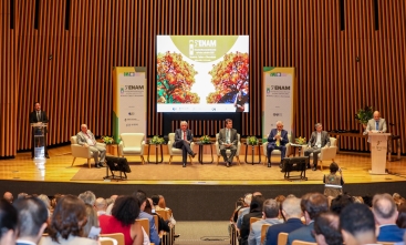 Imagem de um auditório, com um telão, e duas pessoas sentadas sobre o palco