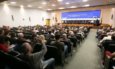 Um evento ocorre em um auditório. No palco, a frente, estão sentadas dez pessoas enquanto uma fala, em pé, em um púlpito. ao fundo, há um telão. Há várias pessoas sentadas em cadeiras pretas enfileiradas voltadas para o palco.