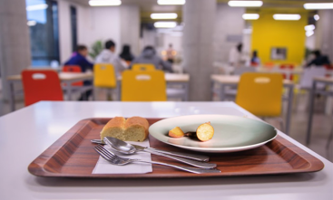 Bandeja sobre a qual há um prato com pouca comida e dois pães pequenos ao lado. Ao fundo, desfocado, um refeitório