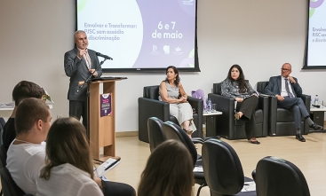 Homem branco, grisalho, de terno cinza e gravata em frente a um púlpito dando uma palestra
