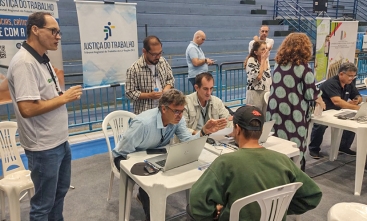 Um homem, trajando um moleton verde escuro e um boné preto está sentado em um posto de atendimento, no qual dois homens fornecem informações. Outros dois homens estão em pé, ao lado da mesa. Ao fundo há dois banners nos quais se lê: Justiça do Trabalho.