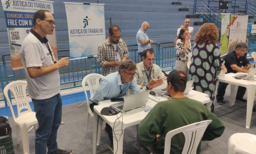 Foto de um atendimento feito por um homem a outro homem, em mesas de plásticos, dentro de um ginásio