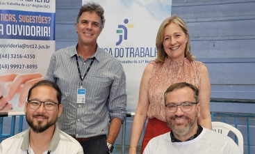 Quatro pessoas sorriem para a foto. Dois homens, sentados; e duas pessoas em pé, sendo um homem e uma mulher. Ao fundo, um banner vertical onde se lê "Justiça do Trabalho"