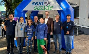 Sete homens e duas mulheres posam em um ambiente aberto, em frente a uma tenda do Sesi. Uma das mulheres está com a canela maquiada, como se tivesse uma fratura exposta